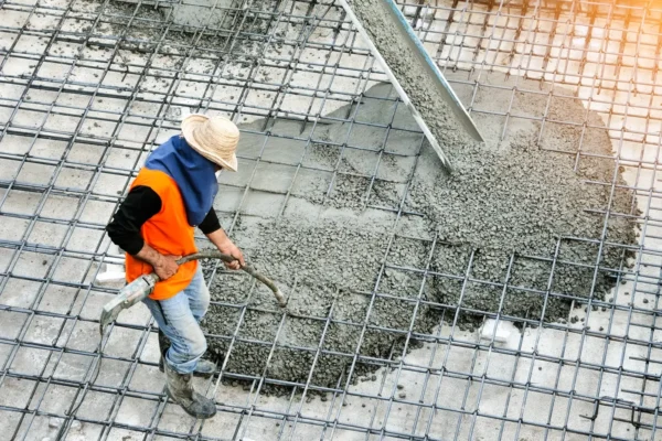 A construction worker wearing an orange vest is pouring concrete onto a concrete floor.