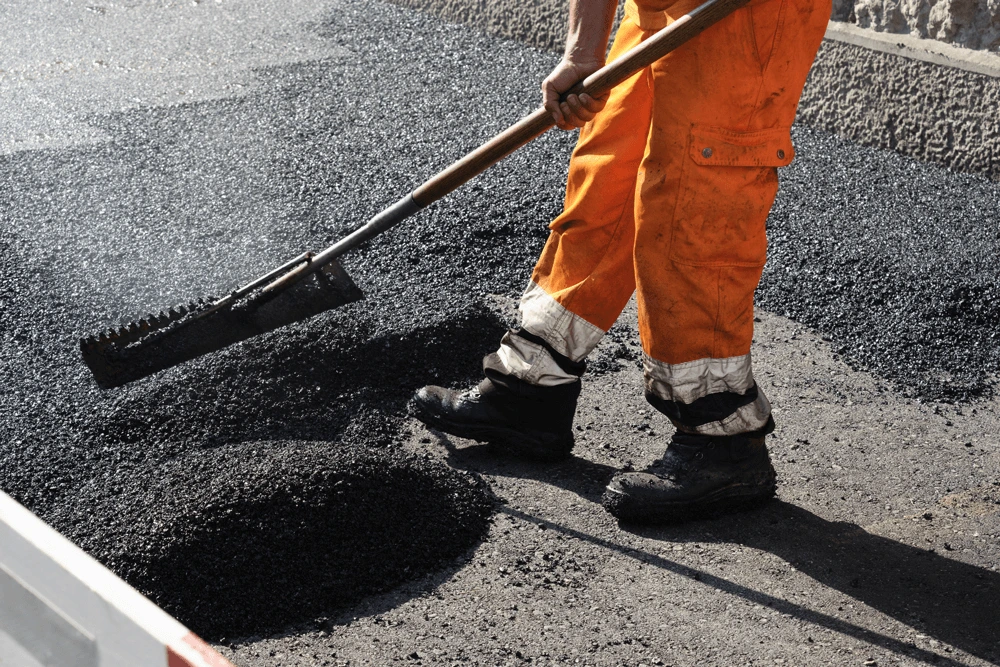 A worker dressed in orange uses a shovel to distribute asphalt on a road construction project.